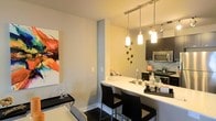 This single-floor space showcases a thoughtfully designed open-concept layout that seamlessly connects the living, dining, and kitchen areas. The kitchen is a standout feature, equipped with dark cabinetry, stainless steel appliances, and a light-colored tile backsplash that adds a clean, modern contrast. A curved breakfast bar with a built-in sink serves as both a functional workspace and a casual dining spot, complemented by sleek black bar stools. Three pendant lights hang above the bar, providing focused task lighting while contributing to the contemporary aesthetic, alongside a track lighting system overhead. The adjacent dining and living area maintains a minimalist character, with neutral-toned walls that create a bright, welcoming atmosphere. A bold, colorful abstract artwork anchors one wall, adding a vibrant artistic accent to the otherwise understated palette. A slim console table with decorative accessories reinforces the space's attention to detail. The overall design balances practicality with modern style, making it a cohesive and inviting environment suited for everyday living and entertaining alike.