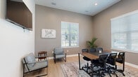 Located at 900 SE Third Ave in Ocala, Florida, this single-floor commercial space functions as a well-appointed business lounge and meeting facility. The layout opens into a bright, airy reception area distinguished by soaring ceilings, light hardwood flooring, and a striking round glass centerpiece table set atop a decorative area rug. Vibrant wall art and a bold accent wall in deep burgundy add a dynamic character to the welcoming entry.

A dedicated conference room offers a comfortable and professional setting for meetings, featuring a large wood-and-metal conference table surrounded by ergonomic task chairs. Multiple windows dressed with white horizontal blinds allow ample natural light to fill the room, while a wall-mounted flat-screen display adds functional versatility. Tasteful wall art, a potted palm, and additional seating along the perimeter complete the space.

A connecting corridor leads to a separate seating nook furnished with modern lounge chairs and a round side table, creating an ideal space for informal conversations or quiet work. Neutral taupe walls, recessed lighting, and clean white trim throughout lend the space a cohesive, professional aesthetic. Signage identifying the "Business Lounge" reinforces the space's professional purpose, making it a practical and inviting environment for work and collaboration.