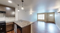 This single-floor unit offers a thoughtfully designed open-concept layout with wood-look flooring running consistently throughout the space. The living area is generously proportioned, featuring neutral-toned walls, a ceiling fan with integrated lighting, and a large window that allows natural light to fill the room. A wall-mounted electrical panel is discretely integrated into the living space.

The kitchen is a standout feature, showcasing rich dark-brown cabinetry with ample upper and lower storage, granite-style countertops, and black appliances including a refrigerator and gas range. A peninsula with a double sink and stylish pendant lighting overhead defines the boundary between the kitchen and living area, creating an efficient and attractive workspace.

The entryway is welcoming, featuring a front door with a window insert that brings in additional natural light. Wall sconces provide ambient lighting throughout. The bathroom is notably accessibility-focused, equipped with stainless steel grab bars near the toilet and bathtub, a roll-in style shower area, and recessed lighting overhead. Additional rooms offer flexible, open spaces with wood-look flooring and neutral finishes, suitable for various uses.