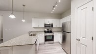 This thoughtfully designed single-floor unit offers a comfortable and functional layout across a well-utilized footprint. The open-concept kitchen and living area serves as the heart of the home, featuring crisp white shaker cabinetry, granite countertops, and a full suite of stainless steel appliances including a range, over-the-range microwave, and refrigerator. A peninsula with an integrated sink provides additional prep space and breakfast bar seating, illuminated by stylish pendant lights and a track lighting system overhead. Light wood-style flooring runs throughout the entire unit, creating a cohesive and modern aesthetic.

The living area is bright and open, with a glass-paneled front door that allows natural light to flow in. A hallway connects the main living space to the private areas of the unit. The spacious bedroom is a generous size, offering a ceiling fan with light fixture, a large window with views of surrounding greenery, and access to a walk-in closet outfitted with wire shelving and hanging rods for ample storage. The bathroom includes a clean, all-white walk-in shower with built-in corner shelving. Additional utility and hallway spaces round out this practical and move-in-ready unit.