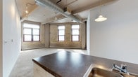This single-floor urban loft showcases a compelling blend of industrial character and modern living. The open-plan layout creates a generous, flexible living area bathed in natural light streaming through large, multi-pane windows framed in deep red wood — a hallmark of classic industrial architecture. The original exposed brick walls in warm yellow tones lend the space a distinctive, historic charm that pairs naturally with the prominent overhead ductwork and concrete ceiling, both hallmarks of a converted industrial building. The kitchen features a substantial island with a dark countertop and an integrated sink, offering both ample prep space and a clear visual anchor for the open living area. Pendant lighting hangs overhead, adding a warm, focused glow to the kitchen zone. The flooring transitions subtly across the space, while ceiling fans contribute to the airy, loft-like atmosphere. The open living area beyond the kitchen is spacious and versatile, with room to accommodate a variety of layout configurations. The combination of raw architectural details, generous window placement, and thoughtful finishes gives this loft a unique, characterful ambiance suited for dynamic urban living.