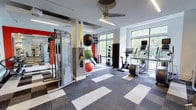 This contemporary fitness center is thoughtfully designed to support a wide range of workout routines. The space features a patterned carpet tile floor in neutral tones of grey and white, providing both comfort and durability underfoot. Large floor-to-ceiling windows line the exterior walls, flooding the interior with abundant natural light and offering views of the surrounding greenery, creating an energizing atmosphere for exercise.

The fitness center is equipped with a comprehensive selection of workout equipment. A multi-functional cable machine serves as a centerpiece for strength training, accompanied by an assortment of stability and medicine balls in various sizes. TRX suspension training straps are conveniently mounted for functional fitness exercises. On the cardio side, an upright stationary bike and an elliptical trainer with integrated digital screens are positioned near the windows to take advantage of the natural light and outdoor views.

A ceiling fan overhead promotes air circulation throughout the space, contributing to a comfortable workout environment. Recessed and linear lighting supplements the natural light for evening use. The clean, open layout ensures ample room to move freely between stations, making this fitness center a versatile and well-appointed facility for residents or members seeking a complete exercise experience.