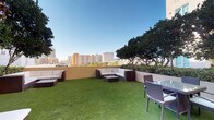 This inviting rooftop terrace offers a well-designed outdoor retreat with a vibrant urban backdrop. The space is covered in lush artificial turf, creating a clean, low-maintenance green surface that lends a park-like feel to the elevated setting. Mature trees line the perimeter, providing natural shade and a touch of greenery against the city skyline. Two distinct seating areas are thoughtfully arranged throughout the terrace. The first features a large L-shaped sectional sofa with neutral cushions framed in dark wicker, paired with a matching coffee table — perfect for group gatherings. The second area offers a glass-top dining table with wicker chairs, suited for outdoor meals or casual conversation. A textured perimeter wall encloses the space, offering both privacy and safety while preserving the open sightlines to the surrounding cityscape. The terrace benefits from abundant natural light and open skies, making it a bright, airy environment throughout the day. The combination of comfortable furnishings, panoramic city views, and thoughtful landscaping makes this rooftop an exceptional space for both relaxation and social entertaining.