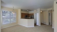 This single-floor residence offers a well-designed open-concept layout that seamlessly connects the kitchen, dining, and living areas, creating a spacious and cohesive living environment. The kitchen is a central highlight, anchored by a wraparound peninsula with granite-style countertops that provide ample prep and seating space. Warm-toned wood cabinetry lines the kitchen walls, complemented by stainless steel appliances including a refrigerator and microwave, as well as track lighting overhead that ensures excellent task illumination. Adjacent to the kitchen, the dining area benefits from a stylish pendant light and a large window fitted with horizontal blinds, allowing generous natural light to fill the space. The living area extends openly from the kitchen and dining zones, featuring neutral wall-to-wall carpeting that adds warmth and comfort underfoot. The soft, neutral wall tones throughout lend the space a clean and inviting ambiance. A hallway visible beyond the kitchen leads to additional rooms, suggesting a practical and well-organized floor plan. Overall, this residence balances functionality with a comfortable, welcoming atmosphere suitable for everyday living.