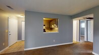 This single-floor residence at 300 Executive Dr in Newark, Ohio, offers a practical and cohesive layout with a neutral gray color palette carried throughout. The kitchen is efficiently designed, featuring gray cabinetry with brushed nickel hardware, granite-look countertops, a double stainless steel sink with under-cabinet lighting, and black appliances including a refrigerator and range. Tile flooring grounds the space and transitions into the adjoining areas.

A distinctive pass-through window connects the kitchen to the main living area, promoting an open feel while maintaining defined zones. The living space is finished with wall-to-wall carpeting and cool gray walls, creating a calm, unified atmosphere. A hallway with tile flooring leads to additional rooms, including a carpeted bedroom with a window allowing natural light and views of the surrounding area.

The bathroom is cleanly appointed with a white vanity, full tub-shower combination with a curtain rod, tile flooring, and gray walls consistent with the rest of the unit. Textured ceilings are present throughout the residence. Overall, the space presents a straightforward, well-maintained interior with consistent finishes across all areas.