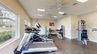 Located at 4793 N Pine Hills Rd in Pine Hills, Florida, this dedicated fitness center offers a well-equipped workout space designed to support a range of exercise routines. The single-room facility is laid out efficiently, making the most of its footprint to accommodate both cardiovascular and strength training equipment. Cardio machines include Spirit-brand treadmills and elliptical trainers, positioned along the windowed wall to allow exercisers to enjoy views of the lush, tree-lined outdoor surroundings while working out. The strength training area features multiple cable and resistance machines, along with a weight bench and additional equipment to support full-body workouts. The interior is finished with neutral beige walls, a crisp white chair rail, and durable concrete flooring. Two ceiling fans provide consistent air circulation throughout the space. Recessed fluorescent lighting ensures the room remains well-lit at all hours. Large windows along one wall flood the space with natural light and frame views of the greenery outside. A colorful piece of wall art adds a vibrant accent to the otherwise understated décor. Multiple entry points, including glass-paneled doors, contribute to the room's accessibility and openness. A wall-mounted hand sanitizer dispenser and a thermostat reflect practical, user-focused amenities.