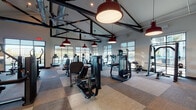 This expansive fitness center is designed to support a comprehensive workout experience within a single, open-plan floor. The space is defined by its striking industrial-inspired architecture, featuring exposed black steel beam trusses, visible ductwork, and bold red pendant lights that hang from the vaulted ceiling — creating a dynamic, energetic atmosphere throughout.

The floor is covered in a patchwork of neutral-toned carpet tiles, providing both durability and visual interest underfoot. Large floor-to-ceiling windows line the perimeter of the space, flooding the interior with natural light and offering views of an outdoor pool area and a waterfront beyond — a particularly scenic backdrop for the treadmill stations positioned directly in front of the water-facing windows.

The fitness center is comprehensively outfitted with a wide selection of Precor and other commercial-grade equipment, including treadmills, ellipticals, cable machines, a Smith machine, weight bench, dumbbell rack, spin bikes, and a dedicated functional training rig complete with a punching bag and medicine balls. A wall-mounted flat-screen television and built-in cabinetry along one wall add convenience and functionality. An oversized ceiling fan aids in air circulation throughout the generous open floor area, completing this well-appointed facility.