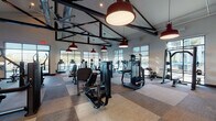 This expansive fitness center is designed to support a comprehensive workout experience within a single, open-plan floor. The space is defined by its striking industrial-inspired architecture, featuring exposed black steel beam trusses, visible ductwork, and bold red pendant lights that hang from the vaulted ceiling — creating a dynamic, energetic atmosphere throughout.

The floor is covered in a patchwork of neutral-toned carpet tiles, providing both durability and visual interest underfoot. Large floor-to-ceiling windows line the perimeter of the space, flooding the interior with natural light and offering views of an outdoor pool area and a waterfront beyond — a particularly scenic backdrop for the treadmill stations positioned directly in front of the water-facing windows.

The fitness center is comprehensively outfitted with a wide selection of Precor and other commercial-grade equipment, including treadmills, ellipticals, cable machines, a Smith machine, weight bench, dumbbell rack, spin bikes, and a dedicated functional training rig complete with a punching bag and medicine balls. A wall-mounted flat-screen television and built-in cabinetry along one wall add convenience and functionality. An oversized ceiling fan aids in air circulation throughout the generous open floor area, completing this well-appointed facility.