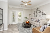 This single-floor residence offers a thoughtfully designed layout that balances comfort and style throughout. The living room sets a welcoming tone with wood-look flooring, crown molding, a ceiling fan, and a large window with access to an outdoor area, allowing natural light to fill the space. Adjacent to the living area, the dining room showcases classic wainscoting panels, soft neutral walls, and elegant crown molding, centered around a round glass-top dining table with seating for four. The kitchen features a galley-style layout with slate-toned cabinetry, light wood-look flooring, a full suite of white appliances including a refrigerator, range, and built-in microwave, along with ample counter and storage space. The bedroom is a serene retreat finished with plush carpeting, a ceiling fan, and large windows dressed in sheer curtains that invite generous natural light. It includes space for a full dresser and nightstands, complemented by abstract wall art. The bathroom continues the home's refined aesthetic with wood-look flooring, a teal-accented vanity, overhead cabinetry, and a clean, functional layout. Throughout the unit, neutral tones, consistent flooring, and quality finishes create a cohesive and inviting ambiance.