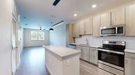 This well-designed 1-bedroom, 1-bathroom unit offers a thoughtful layout across a single floor, combining modern finishes with functional living spaces. The open-concept kitchen serves as the centerpiece of the home, featuring light wood cabinetry extending to the ceiling for ample storage, stainless steel appliances including a French door refrigerator, dishwasher, and range, as well as a granite-topped island that provides additional prep space and seating. Recessed lighting and a sleek linear pendant fixture illuminate the kitchen, which flows seamlessly into the living area. The living room is bright and airy, benefiting from large windows that invite natural light and offer outdoor views. Light wood-style plank flooring runs throughout the main living areas, lending a clean, contemporary feel. The bedroom is a comfortable retreat with soft carpeting, two generously sized windows with outdoor views, and access to a walk-in closet. The bathroom is finished with matching granite countertops, a wood-toned vanity with under-sink storage, a framed mirror, and tile flooring. A hallway connects the rooms efficiently, completing this cohesive and modern living space.