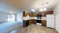 This single-floor home offers a well-designed layout that balances practicality and comfort. The kitchen serves as a central hub, featuring warm-toned wooden cabinetry with decorative raised-panel detailing that provides generous storage throughout. The countertops extend across an L-shaped configuration, offering ample workspace for meal preparation. A double-basin sink is positioned beneath overhead cabinetry, while black appliances — including a built-in microwave and dishwasher — contrast neatly with a white electric range. The tile flooring in the kitchen adds durability and visual structure to the space. An elegant chandelier illuminates the dining area adjacent to the kitchen, complemented by a fluorescent ceiling fixture above the main work area. A textured popcorn ceiling runs throughout, adding a classic residential character. The kitchen flows openly into a carpeted living area, which benefits from abundant natural light streaming through a large window or sliding door at the far end — creating a bright and welcoming atmosphere. The seamless connection between the kitchen and living area enhances the overall sense of openness, making this home well-suited for everyday living and casual entertaining.