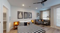 This single-floor studio unit offers a well-organized open-concept layout that efficiently combines living, sleeping, and dining functions within one cohesive space. Light wood-tone flooring runs throughout, creating a warm, unified foundation that ties each area together seamlessly.

The sleeping and living area occupies the main portion of the space, where a full-size bed with a warm wood headboard platform sits alongside a gray sectional sofa, defining distinct zones without physical dividers. A ceiling fan with recessed lighting overhead provides both comfort and illumination. Botanical fern artwork on soft gray walls adds a consistent decorative motif, and industrial-style pipe shelving near a window with sheer drapes offers functional display space. Natural light filters in through the window, brightening the area.

The kitchen is clean and contemporary, featuring white flat-panel cabinetry, light quartz countertops, a tile backsplash, and a full suite of stainless steel appliances including a refrigerator, range, microwave, and dishwasher. A compact butcher-block dining table with industrial stools sits adjacent, providing a casual dining option.

The bathroom includes a combination tub and shower with tiled surround, recessed lighting, and wood-look flooring consistent with the rest of the unit, maintaining a cohesive aesthetic throughout.