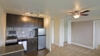 This single-floor studio features an open-concept layout that efficiently combines the kitchen and living areas into one cohesive space. The kitchen is appointed with dark espresso cabinetry, stainless steel appliances including a full-sized refrigerator and dishwasher, and a mosaic tile backsplash that adds a decorative accent. A stainless steel sink and granite-style countertop complete the culinary space, with a track lighting fixture overhead providing focused illumination. The transition from the kitchen's hardwood-style flooring to the carpeted living area delineates the two functional zones. The living space is equipped with a ceiling fan with integrated light fixture, offering both comfort and ambient lighting. Two built-in closets with bi-fold doors provide ample storage, one of which features visible shelving and a hanging rod. The neutral wall tones and light-colored carpet contribute to a clean, understated aesthetic throughout the space.