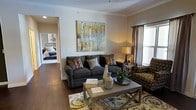 This single-floor residence offers a thoughtfully designed layout that balances comfort and style. The living area serves as the heart of the home, featuring rich hardwood flooring, crisp crown molding, and neutral-toned walls that create a bright and inviting atmosphere. A large statement artwork depicting birch trees anchors the main wall, adding a natural, artistic focal point to the room. The space is furnished with a plush, charcoal sofa accented by colorful throw pillows, complemented by a patterned accent chair that adds visual depth. A rectangular coffee table with clean lines sits atop a floral area rug, bringing warmth and texture to the floor. A classic table lamp provides soft ambient lighting, while a window dressed with layered curtains allows for natural light control. The open sightline from the living area extends directly into the bedroom, which features a wrought-iron bed frame and continues the same hardwood flooring, creating a cohesive aesthetic throughout. The overall ambiance of this residence is cozy yet refined, with a curated mix of traditional and transitional design elements that make it feel both livable and well-appointed.