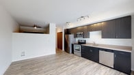 This single-floor space showcases a thoughtfully designed open-concept layout that seamlessly connects the kitchen and living areas. The kitchen is a standout feature, equipped with a full suite of stainless steel appliances including a French door refrigerator, electric range with oven, built-in microwave, and dishwasher. Rich, dark-toned cabinetry lines the walls, providing ample storage, while a clean countertop with an undermount sink adds to the contemporary aesthetic. Track lighting installed on the ceiling ensures bright, focused illumination throughout the kitchen workspace. The flooring throughout the unit consists of light-toned, wood-style planks that add warmth and continuity to the space. White walls and a clean white ceiling contribute to an open, airy ambiance, while the raised ceiling in the living area adds a sense of volume. A partial wall divider subtly separates the living space from the kitchen zone without compromising the open feel. A ceiling fan is visible in the adjoining living area, enhancing comfort. A wood-toned door leads to additional rooms or storage, completing this well-appointed, modern single-floor space.