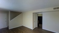 This two-floor residence offers a practical and functional layout designed for comfortable everyday living. The main level showcases rich, dark hardwood-style flooring that extends throughout the open living area, creating a warm and inviting atmosphere. A staircase positioned along one wall leads to the upper floor, making efficient use of the available space while adding a structural focal point to the room. The living area is finished with neutral, light beige walls that lend a clean and versatile backdrop, complemented by crisp white trim and baseboards. The textured popcorn ceiling adds a classic touch overhead. An open doorway from the main living space provides direct access to the kitchen, which is visible in the background and equipped with dark appliances. The overall layout is straightforward and well-connected, allowing for easy movement between the living and kitchen areas. The combination of dark flooring, neutral walls, and white accents gives the space a balanced, understated aesthetic suitable for a variety of interior styles and furnishing choices.