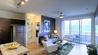 This single-floor residence showcases a thoughtfully designed open-concept layout that seamlessly connects the living and kitchen areas. The living room serves as the centerpiece of the space, highlighted by a bold dark blue accent wall adorned with decorative artwork, creating a striking focal point against the otherwise neutral color palette of soft whites and warm grays.

Natural light floods the interior through expansive floor-to-ceiling sliding glass doors, which open to a private balcony offering outdoor views of lush greenery. A ceiling fan with an integrated light fixture enhances comfort and airflow throughout the space. The living area comfortably accommodates a sectional sofa, accent chair, and a contemporary coffee table atop a patterned area rug, all resting on warm hardwood-style flooring.

An ornate mirrored room divider elegantly separates the living space from the kitchen area, where stainless steel appliances, including a full-size refrigerator, are visible. A hallway beyond the divider leads to additional rooms. The overall ambiance is modern and stylish, balancing comfort with contemporary design elements.