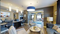 This single-floor space showcases a thoughtfully designed open-plan layout that seamlessly connects multiple functional zones. Bold cobalt-blue drum pendant lights serve as striking focal points, anchoring each area while maintaining visual continuity throughout the space. The color palette pairs warm taupe and charcoal accent walls with soft yellow tones, creating a dynamic yet cohesive aesthetic.

A dedicated business center is positioned along one side, featuring a built-in granite-topped desk with dual computer workstations, ergonomic seating, and ample task lighting — ideal for productivity. Adjacent to this, a casual lounge area is furnished with patterned accent chairs and a metallic round coffee table, offering a relaxed seating option.

Through an open archway, a secondary lounge area is visible, featuring natural light from large windows, additional seating arrangements in neutral tones, and vibrant accent pieces including orange throw pillows and a blue area rug. Hardwood-style flooring flows throughout, unifying the entire space and adding warmth. The overall ambiance is modern and welcoming, balancing professional amenities with comfortable social gathering areas.