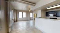 This single-floor loft-style space showcases an open-concept layout that seamlessly blends industrial character with comfortable living. The expansive main living area is anchored by polished concrete floors that lend a sleek, modern aesthetic, while exposed structural beams running across the ceiling add a distinctive industrial charm. A ceiling fan with an integrated light fixture provides both function and a classic finishing touch overhead.

Natural light pours in through a row of large, evenly spaced windows along one wall, fitted with blinds for adjustable privacy. The generous ceiling height enhances the sense of openness and volume throughout the space.

The kitchen area is partially separated by a white half-wall peninsula, topped with a light granite or stone-style counter surface, offering a natural division between the living and culinary spaces while maintaining the open flow. A dark refrigerator is visible at the kitchen's edge, hinting at a well-appointed cooking area beyond.

Toward the rear of the space, a doorway leads to what appears to be a walk-in closet or additional room, providing practical storage solutions. Exposed piping along the upper walls and ceiling further reinforce the loft's raw, urban character, making this a distinctive and versatile space well-suited to a variety of uses.