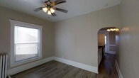 This single-story space offers a clean, functional layout with a warm and inviting aesthetic. The main living area is finished with rich, dark hardwood-style flooring that runs throughout, providing a cohesive and grounded feel. Neutral beige walls create a versatile backdrop, complemented by crisp white baseboards that add a classic finishing touch. A ceiling fan with a built-in light fixture is centrally mounted, offering both comfort and practical illumination. Natural light enters through a double-hung window fitted with blinds, brightening the space during the day. A standout architectural detail is the elegant arched doorway that connects the living area to the kitchen, adding a touch of traditional character to the overall design. The kitchen, visible through the arch, features cabinetry along the walls and access to a rear entry door with a window, allowing for additional natural light. The layout flows intuitively, making the space feel open and well-connected. Overall, the design balances simplicity with timeless architectural details, creating a comfortable and practical environment suited for everyday living.