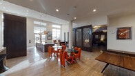 This thoughtfully designed single-floor amenity space combines bold contemporary style with a variety of functional areas, creating an engaging environment for socializing and recreation. The main lounge entrance greets visitors with soaring ceilings, floor-to-ceiling windows that flood the space with natural light, and polished tile flooring that reflects the bright, open atmosphere. Vibrant orange sculptural chairs surround a sleek bistro table, adding a striking pop of color against the neutral palette.

A sophisticated bar and kitchen area features a large island with upholstered counter seating, an eye-catching arrangement of globe pendant lights in amber and chrome, and a textured metallic backsplash that anchors the space with contemporary flair. Nearby, a row of minimalist pedestal bar stools lines a secondary counter beneath a decorative honeycomb partition.

The lounge area is defined by dramatic dark-wood honeycomb screen panels that enclose a comfortable seating nook with a deep sectional leather sofa, a sculptural organic coffee table, and an elegant floral chandelier overhead. An additional lounge nook features moody charcoal walls adorned with a gallery of framed artwork and plush leather seating.

Completing the space is a dedicated indoor golf simulator room, fitted with turf flooring, projection equipment, and a large screen, offering a unique recreational amenity within this impressively curated environment.
