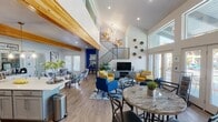 This vibrant two-story space is thoughtfully designed to accommodate a wide variety of activities and functions. The main level opens into a dramatic, soaring great room with vaulted ceilings, exposed natural wood beams, and light wood flooring throughout. A striking open staircase with metal railings connects the two floors, while large glass doors and expansive windows flood the space with natural light and provide direct views of an outdoor pool area.

The fully equipped kitchen is a standout feature, boasting grey shaker-style cabinetry, stainless steel appliances, a mosaic tile backsplash, a large central island with bar seating, and bold pendant lighting beneath the wood beam ceiling. Adjacent to the kitchen, a dining and lounge area offers a marble-top table and a comfortable seating arrangement with colorful accent chairs.

A dedicated yoga and fitness studio offers an open, serene environment complete with exercise balls, yoga mats, wall mirrors, and motivational artwork. The children's activity rooms are bright and imaginative, featuring small-scale tables and chairs, a large built-in bookshelf with colorful cubbies, whiteboards, and ample storage. A quiet workspace nook with pool-facing French doors rounds out this well-appointed, multi-functional space.