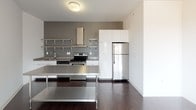 This contemporary two-bedroom, two-bathroom Dwell unit is designed with a clean, modern aesthetic throughout its single floor layout. The open-concept living and kitchen area serves as the heart of the home, featuring rich dark hardwood floors that contrast beautifully with the bright white walls. Large windows along the living area flood the space with natural light while offering pleasant views of the surrounding neighborhood rooftops and tree canopy.

The kitchen is thoughtfully designed with stainless steel open shelving, a stainless steel electric range with a prominent ventilation hood, a stainless steel refrigerator, a deep sink, and sleek white lower cabinetry topped with gray countertops. The industrial-modern aesthetic is further emphasized by a stainless steel work surface in the kitchen area.

Both bedrooms are finished with plush neutral carpeting and crisp white walls, creating calm, restful retreats. Each bedroom benefits from window light, with one featuring direct neighborhood views through a large dark-framed window.

The two bathrooms share a consistent design language, each featuring warm terracotta-toned tile flooring, white walls, a dark wood vanity with a white countertop and integrated sink, and a towel bar. The overall ambiance of this unit is clean, contemporary, and move-in ready.