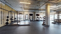 This well-equipped fitness center showcases a striking industrial aesthetic defined by exposed concrete ceilings, visible ductwork, and structural columns that lend the space a raw, urban character. The layout is generously proportioned, providing ample room for a diverse range of workout activities without feeling crowded.

A dedicated strength training zone features a power rack, barbell platforms with wooden lifting surfaces, and a full dumbbell rack, set against a distinctive yellow corrugated metal wall bearing the facility's branding. This area provides a functional and visually bold backdrop for resistance training.

The cardio section is positioned along a windowed perimeter wall, where multiple treadmills, elliptical trainers, and a StairMaster benefit from natural light and exterior views. Wall-mounted flat-screen televisions are positioned throughout the space for entertainment during workouts.

Additional equipment includes medicine ball racks, a cable machine, and a punching bag, supporting functional and cross-training exercises. The entire floor is covered in durable rubber flooring, ensuring safety and comfort. Large garage-style windows and glass doors further brighten the space, creating an open, energetic atmosphere well-suited to a full range of fitness activities.