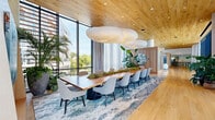 Located at 4400 Calvert Rd in College Park, Maryland, this thoughtfully designed single-floor amenity space seamlessly blends natural materials with modern aesthetics to create an inviting and functional environment. Throughout the space, warm wood-planked ceilings and light hardwood flooring establish a cohesive organic palette that extends from one end to the other.

A defining architectural feature is the expansive wall of floor-to-ceiling louvered windows lining the length of the space, allowing abundant natural light to filter in while offering dynamic views of the surrounding landscape and neighboring buildings. Sheer white curtains complement the louvers, providing a soft, airy quality to the interiors.

The space is organized into distinct yet open zones. A generous lounge area features plush seating arrangements — including deep sofas, upholstered armchairs in warm cognac leather, and accent chairs — positioned atop patterned area rugs, creating an atmosphere ideal for relaxed gatherings. Lush tropical plants are thoughtfully placed throughout, reinforcing the biophilic design philosophy.

A striking conference or dining area anchors one end of the space, centered around a live-edge wood table surrounded by soft grey upholstered chairs. Sculptural cloud-like pendant lights float above, adding an artistic focal point. Frosted glass-paneled private rooms provide additional enclosed spaces along the interior wall, completing this refined and welcoming communal environment.