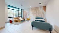 Located at 1559 Northwest 15th Street Road in Miami, FL, this single-floor space offers a well-appointed layout comprising a living room and a kitchen. The living room is a standout area, characterized by large floor-to-ceiling windows with sleek black frames that invite abundant natural light and provide open views of the surrounding greenery. The room features light-toned large-format tile flooring throughout, recessed lighting, and a striking feature wall adorned with an artistic botanical mural — a bold design element that adds a vibrant, tropical personality to the space. Comfortable seating arrangements are complemented by a warm neutral color palette and contemporary furnishings, including accent chairs in mustard yellow and teal tones that echo Miami's lively aesthetic. The kitchen is equally refined, showcasing floor-to-ceiling cabinetry in a warm greige finish with matte black hardware, a crisp white countertop, and an eye-catching teal herringbone tile backsplash that serves as a vivid focal point. A stainless steel French door refrigerator adds a polished, modern touch. The space also features a louvered interior door, adding a functional architectural detail. Overall, the space presents a clean, contemporary design with thoughtful color accents and a bright, welcoming ambiance.