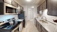 This single-floor residence offers a thoughtfully designed layout with a clean, modern aesthetic throughout. The galley-style kitchen is a standout feature, equipped with stainless steel appliances including a refrigerator, electric range, and built-in microwave. Dark espresso cabinetry lines both walls, complemented by granite-style countertops and a stainless steel sink, all set atop light wood-style luxury vinyl flooring that extends through the main living areas.

The bathroom continues the cohesive design language, featuring a matching dark vanity with granite countertops, a framed mirror, and contemporary brushed nickel fixtures. An adjacent water closet provides added privacy and functionality. The bedroom areas are finished with plush neutral carpeting, offering a comfortable contrast to the hard-surface flooring elsewhere. One bedroom includes a walk-in closet outfitted with dual wire shelving rods, providing generous storage capacity.

Additional highlights include baseboard heating, a wall-mounted HVAC unit, and textured ceilings in select areas. The rooms are bright and neutral in tone, with white walls that maximize the sense of openness. Windows with blinds allow for natural light while maintaining privacy, contributing to the overall clean and functional character of the space.