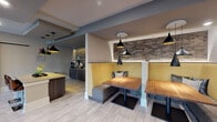 Located at 900 Hobbs Rd in Greensboro, NC, this single-floor community clubhouse and leasing office blends contemporary design with warm, inviting elements. Light-toned tile flooring flows throughout the open layout, complementing the neutral gray walls and creating a bright, welcoming atmosphere.

The lounge and café area serves as the heart of the space, anchored by a Starbucks coffee station set against a striking textured stone accent wall. A central island with a quartz-style countertop and mid-century modern bar stools provides a casual gathering spot, while sleek dark cabinetry and a compact refrigerator complete the beverage station. Overhead, a cluster of stylish black pendant lights adds a modern industrial touch.

Adjacent booth seating features mustard-yellow upholstered banquettes, natural wood tabletops, and additional pendant lighting — all framed by the same stone accent wall, creating semi-private nooks ideal for conversation or relaxation.

A dedicated workstation area offers a desktop computer and task-oriented desk, accented by a sculptural branch-style light fixture and greenery, with a large glass door providing views to an outdoor patio area. Dramatic dark-stained barn-style sliding doors serve as a bold architectural focal point, adding rustic contrast to the otherwise modern interior. Glass partition walls near the entrance enhance openness and allow natural light to filter throughout the space.