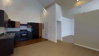 This one-bedroom, one-bathroom residence is designed to maximize comfort and functionality within a well-proportioned floor plan. The space immediately impresses with its dramatic vaulted ceilings, which lend an expansive, airy quality to the interior and enhance the sense of openness throughout. The kitchen is outfitted with dark cabinetry featuring warm wood accents, black appliances including a full-size refrigerator and gas range, and hardwood-style flooring that grounds the cooking area with a rich, contrasting finish. The kitchen transitions seamlessly into the main living area, which is finished with neutral carpet that adds warmth and comfort underfoot. An open archway connects the living space to the bedroom, maintaining the home's cohesive, flowing layout. White walls throughout keep the interior bright and clean, complementing the high ceilings to amplify natural light. The overall design is straightforward and practical, offering a move-in-ready environment that balances everyday living needs with architectural interest — most notably the striking vaulted ceiling that sets this residence apart from standard single-story layouts.