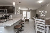 This single-floor residence offers a thoughtfully designed open-concept layout that seamlessly connects the kitchen, dining, and living areas. The kitchen is a standout feature, equipped with dark espresso cabinetry, stainless steel appliances including a gas range, microwave, and refrigerator, complemented by light granite countertops and a classic subway tile backsplash. A peninsula with bar-height seating creates a natural transition into the living space, making it ideal for both everyday living and entertaining. The living room is bright and welcoming, with neutral taupe walls, plush carpeting, a ceiling fan, and a glass door that invites natural light and provides outdoor access. A dedicated dining nook accommodates a round table and chairs, offering a cozy space for meals. The bedroom is a serene retreat with soft carpet, a ceiling fan, and large windows dressed in sheer curtains that allow ample natural light. The bathroom features tile flooring, a granite-topped dark wood vanity, and a shower-tub combination. Throughout the space, a cohesive neutral palette and consistent dark wood accents tie each area together, creating a harmonious and comfortable living environment.