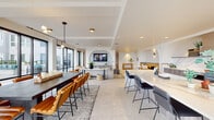 This thoughtfully designed single-floor amenity space offers a sophisticated blend of modern architecture and comfortable social living. Anchored by a striking open-plan layout, the space flows seamlessly between distinct functional zones connected by elegant arched architectural details that lend warmth and character throughout.

The fully equipped kitchen serves as a central hub, featuring a dramatic marble-look quartz island with bar seating, custom wood cabinetry, a bold blue professional-grade range, a marble backsplash, a beverage cooler, and brass-accented fixtures. Large-format stone tile flooring extends throughout, adding a cohesive, polished aesthetic.

The dining area accommodates generous group gatherings around a substantial dark-toned table with cognac leather chairs, all beneath a contemporary linear pendant light. Floor-to-ceiling black-framed glass doors open directly to an outdoor courtyard, flooding the space with natural light and providing seamless indoor-outdoor connectivity.

The lounge area offers multiple comfortable seating arrangements, including a large sectional sofa and accent chairs, centered around a marble coffee table. A feature arched alcove houses a large-screen television above custom cabinetry. A built-in banquette with vibrant yellow upholstery adds a pop of color to a more intimate nook, while a billiards table rounds out the entertainment offerings.