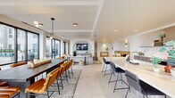 This thoughtfully designed single-floor amenity space offers a sophisticated blend of modern architecture and comfortable social living. Anchored by a striking open-plan layout, the space flows seamlessly between distinct functional zones connected by elegant arched architectural details that lend warmth and character throughout.

The fully equipped kitchen serves as a central hub, featuring a dramatic marble-look quartz island with bar seating, custom wood cabinetry, a bold blue professional-grade range, a marble backsplash, a beverage cooler, and brass-accented fixtures. Large-format stone tile flooring extends throughout, adding a cohesive, polished aesthetic.

The dining area accommodates generous group gatherings around a substantial dark-toned table with cognac leather chairs, all beneath a contemporary linear pendant light. Floor-to-ceiling black-framed glass doors open directly to an outdoor courtyard, flooding the space with natural light and providing seamless indoor-outdoor connectivity.

The lounge area offers multiple comfortable seating arrangements, including a large sectional sofa and accent chairs, centered around a marble coffee table. A feature arched alcove houses a large-screen television above custom cabinetry. A built-in banquette with vibrant yellow upholstery adds a pop of color to a more intimate nook, while a billiards table rounds out the entertainment offerings.