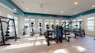 This fitness center is a thoughtfully designed, single-floor facility that combines functionality with a clean, modern aesthetic. The space is defined by its striking teal accent beams and trim that contrast sharply against crisp white walls and ceilings, creating a visually cohesive and energizing environment. Large multi-pane windows line the walls on multiple sides, flooding the interior with abundant natural light and offering views of lush outdoor greenery and a nearby pool area.

The flooring throughout is a light hardwood-style material that enhances the bright, open feel of the space. Recessed lighting and ceiling fans supplement the natural light, ensuring comfort during any time of day.

The fitness floor is comprehensively equipped with a wide range of cardio machines, including stationary bikes, ellipticals, and treadmills with individual screens, all positioned to take advantage of the natural light and outdoor views. Strength training is well-represented with a multi-station cable machine, a Smith machine with a bench, a dedicated free weights area with a tiered dumbbell rack, and a weight bench.

A convenience area near the exit features teal-painted cubbies for personal storage, a mini-refrigerator, and a wall-mounted flat-screen television. Additional wall-mounted TVs are positioned throughout the space to keep members entertained during workouts. The space is clean, open, and purposefully organized for an effective fitness experience.