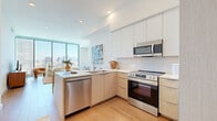 Located on the 21st floor of a high-rise building along Wilshire Boulevard in Los Angeles, this one-bedroom, one-bathroom residence offers a sleek, modern aesthetic with an open and efficient layout. Light wood flooring flows throughout, complementing the clean white walls and contemporary design.

The open-plan living, dining, and kitchen area forms the heart of the home. The kitchen is outfitted with flat-front cabinetry in white and warm wood tones, stainless steel appliances including a range, microwave, dishwasher, and refrigerator, and a peninsula with bar seating. The adjacent dining area provides space for a full table, while the living room is anchored by dramatic floor-to-ceiling corner windows that frame sweeping panoramic views of the Los Angeles skyline and surrounding cityscape, flooding the space with natural light.

The bedroom continues the floor-to-ceiling window treatment, offering the same breathtaking city views and abundant daylight. A dedicated walk-in closet provides well-organized storage with built-in shelving and hanging rods.

The bathroom features large-format stone-look tile flooring, a wall-mounted toilet, and a full glass-enclosed walk-in shower, lending a clean and spa-like feel. The overall ambiance of the unit is bright, airy, and distinctly urban, with modern finishes and elevated high-rise living at its core.