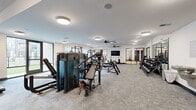 Located at 6163 Kempsville Circle in Norfolk, Virginia, this generously sized fitness facility spans a single floor and is divided into two distinct exercise rooms, each thoughtfully designed to support a full range of training needs. The main strength training room features plush carpet flooring, large ceiling fans, and abundant recessed and flush-mount lighting that keeps the space bright and energizing. This area is equipped with a comprehensive array of machines, including cable systems, multi-use benches, a full dumbbell rack, rowing machines, and cardio equipment such as ellipticals and air bikes. A wall-mounted flat-screen TV adds a modern touch, while large windows invite natural light and outdoor views. Mirrored walls with sleek black-framed glass partitions visually expand the space and provide connectivity between zones. The second exercise room features warm hardwood flooring and is purpose-built for functional and high-intensity training. A full-size glass garage door opens to provide fresh air and an airy feel. This room houses kettlebells, battle ropes, resistance bands, plyo boxes, a tire flip station, a power rack, and a TRX suspension system. Large ceiling fans maintain airflow throughout both rooms. A welcoming entry area with carpet, a water fountain, and sanitation station completes this well-appointed fitness center.
