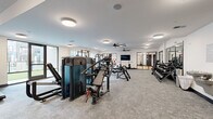 Located at 6163 Kempsville Circle in Norfolk, Virginia, this generously sized fitness facility spans a single floor and is divided into two distinct exercise rooms, each thoughtfully designed to support a full range of training needs. The main strength training room features plush carpet flooring, large ceiling fans, and abundant recessed and flush-mount lighting that keeps the space bright and energizing. This area is equipped with a comprehensive array of machines, including cable systems, multi-use benches, a full dumbbell rack, rowing machines, and cardio equipment such as ellipticals and air bikes. A wall-mounted flat-screen TV adds a modern touch, while large windows invite natural light and outdoor views. Mirrored walls with sleek black-framed glass partitions visually expand the space and provide connectivity between zones. The second exercise room features warm hardwood flooring and is purpose-built for functional and high-intensity training. A full-size glass garage door opens to provide fresh air and an airy feel. This room houses kettlebells, battle ropes, resistance bands, plyo boxes, a tire flip station, a power rack, and a TRX suspension system. Large ceiling fans maintain airflow throughout both rooms. A welcoming entry area with carpet, a water fountain, and sanitation station completes this well-appointed fitness center.