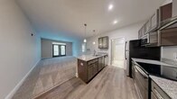 Located at 130 Canary Place in Burlington, NC, this 1-bedroom, 1-bathroom residence offers a thoughtfully designed single-floor layout with modern finishes throughout. The open-concept kitchen and living area serves as the heart of the home, anchored by a center island with granite countertops, an undermount sink, and elegant pendant lighting. Dark-toned shaker-style cabinetry with glass-front upper cabinets complements the full suite of stainless steel appliances, including a range with a wall-mounted hood vent, refrigerator, microwave, and dishwasher. Wood-look plank flooring runs through the kitchen, transitioning to plush carpet in the spacious living room, which features French doors that open to a private outdoor balcony, allowing natural light to fill the space. The bedroom is comfortably sized, fitted with soft carpet, a ceiling fan, and windows overlooking lush greenery. The bathroom includes a full tub-shower combination, a granite-topped vanity, and tile flooring. A dedicated utility room completes the layout. Neutral gray tones and clean white trim throughout lend the space a fresh, contemporary aesthetic.