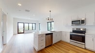 This contemporary single-floor residence showcases a thoughtfully designed open-concept layout, seamlessly connecting the kitchen and living area in a bright, airy environment. Warm hardwood-style flooring flows consistently throughout the entire space, complementing the clean white walls and modern aesthetic.

The kitchen is a standout feature, centered around a peninsula with a built-in sink and stainless steel dishwasher. White shaker-style cabinetry extends to the ceiling, paired with quartz countertops and a white subway tile backsplash. Stainless steel appliances — including an electric range, over-the-range microwave, and refrigerator — complete the well-appointed cooking space. A striking multi-bulb chandelier anchors the area, adding a touch of industrial-modern style. Large black-framed windows and sliding glass doors flood the open living space with abundant natural light and outdoor views.

The space includes two bathrooms: one featuring a walk-in glass-enclosed shower with large-format tile and an LED-backlit vanity mirror, and a second with a full soaking tub and shower combination. Bedrooms are bright and airy, with large windows offering leafy outdoor views. A generously sized walk-in closet with wire shelving provides ample storage. The overall ambiance is clean, modern, and move-in ready.