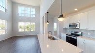This two-story residence is designed with a bright, open layout and modern finishes throughout. The main level centers around a dramatic living area with soaring two-story ceilings and an expansive wall of windows, flooding the space with natural light and offering views of surrounding greenery. Dark hardwood-style flooring anchors the main living areas, creating a clean contrast against the neutral gray walls.

The kitchen is well-appointed with white shaker cabinetry, light gray subway tile backsplash, quartz countertops, and a large center island with an undermount sink. Stainless steel appliances—including a range, microwave, and refrigerator—complete the functional cooking space. Pendant lighting above the island adds a contemporary touch.

The upper level features carpeted bedrooms, each with ceiling fans and windows overlooking the outdoors. One bedroom offers direct access to a bathroom equipped with a walk-in closet and a neo-angle glass shower. A second full bathroom includes a tub-shower combination with built-in shelving. An additional carpeted bedroom with an adjacent bathroom and walk-in closet provides flexible living arrangements. The overall aesthetic is clean, modern, and move-in ready.