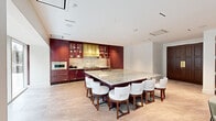 This sophisticated single-floor space is designed for refined entertaining, seamlessly blending a gourmet kitchen, casual dining lounge, and formal dining room into one cohesive layout. The chef's kitchen is a standout centerpiece, featuring deep burgundy cabinetry with gold hardware, a striking brass range hood, marble slab backsplash, and professional-grade stainless steel appliances. A generously proportioned marble-topped island with seating for multiple guests anchors the space and flows naturally into the casual dining and lounge area, which includes built-in banquette seating, a wall-mounted display screen, and an oval dining table with upholstered chairs. The formal dining room exudes drama and sophistication, adorned with richly detailed floor-to-ceiling botanical wallcoverings, a statement chandelier, a long dark dining table with teal velvet chairs, and large sliding glass doors that open directly onto a landscaped outdoor terrace with manicured greenery and wooden privacy screens. A connecting corridor features dark green lacquered built-in cabinetry and ornate display shelving, adding an intimate and curated touch. Throughout, polished marble-style tile flooring and recessed lighting contribute to the space's luxurious, gallery-like ambiance.