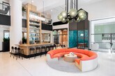 Located at 1901 E Dyer Rd in Santa Ana, California, this single-floor commercial space delivers a striking blend of contemporary design and vibrant artistic expression. The grand lobby anchors the layout with soaring double-height ceilings, a sweeping circular orange sofa beneath an eye-catching globe chandelier, and a built-in bar and shelving area branded "The Goods," creating an immediate sense of energy and sophistication. A textured sculptural wall adds architectural depth to the main gathering area, while warm wood slat accents and polished white floors maintain a refined aesthetic throughout. Multiple distinct lounge zones offer flexible seating configurations, including a relaxed area with navy sectional sofas and framed interior windows, and a sunlit nook furnished with rattan egg chairs and teal cushions set against vibrant wall art. Floor-to-ceiling glass walls and large windows invite abundant natural light and outdoor views of surrounding greenery. A glass-enclosed conference room, wrapped in teal marbled paneling, provides a visually dramatic private meeting space. The fully equipped galley-style kitchen features white quartz countertops, wood cabinetry, a gas cooktop, and a slatted wood ceiling, complemented by a spacious island kitchen with bar seating and distinctive artistic wall pieces. The overall ambiance is creative, welcoming, and professionally curated.