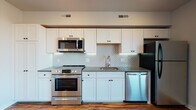 This well-appointed single-floor unit showcases a thoughtfully designed open-concept layout that seamlessly blends the kitchen and living areas. The kitchen is a standout feature, outfitted with crisp white shaker-style cabinetry, gray countertops, and a striking herringbone-patterned tile backsplash. A full suite of stainless steel appliances — including a range, over-the-range microwave, dishwasher, and refrigerator — adds a polished, modern touch. Rich hardwood-style flooring runs throughout the main living area, complementing the neutral walls and recessed lighting overhead. A large picture window in the living space frames expansive urban rooftop views, flooding the room with natural light. The bedroom offers plush carpeting underfoot, a built-in closet, and a calm, neutral palette ideal for rest and relaxation. The bathroom is finished with beige tile flooring, clean white walls, and a glass-enclosed walk-in shower adorned with marble-look wall panels and matte black hardware accents. Additional HVAC vents throughout ensure year-round comfort. Overall, the space presents a clean, contemporary aesthetic with quality finishes and functional design at every turn.
