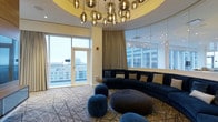 This single-floor lounge space showcases a refined contemporary design, highlighted by a striking circular ceiling recess adorned with a cascading chandelier of amber-toned glass globe pendants that serve as an immediate focal point upon entry. The open-plan layout is anchored by expansive floor-to-ceiling windows and glass partitions on multiple sides, flooding the interior with abundant natural light while offering sweeping elevated views of the surrounding cityscape. A set of glazed doors provides direct access to an outdoor area, further connecting the interior to its urban setting. The flooring features a geometric-patterned carpet in cool grey tones, complementing the space's sophisticated color palette. Seating is centered around a large curved navy-blue sectional sofa, accented with gold and patterned cushions, and accompanied by organic-shaped velvet ottomans in deep blue. Floor-length beige drapery softens the window walls, adding warmth to the otherwise sleek aesthetic. The overall ambiance is luxurious yet approachable, blending modern architectural details with plush, carefully curated furnishings suited to an upscale lounge or hospitality environment.