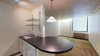 This single-floor unit offers a practical and well-organized layout designed to maximize functionality. The galley-style kitchen is equipped with white cabinetry, a refrigerator, electric range, microwave, and dark laminate countertops. A classic black-and-white checkered tile floor adds a distinctive visual element, while wall-mounted wire shelving provides additional storage. The kitchen opens to the living area via a curved peninsula counter topped with a pendant light, creating a natural division between the two spaces. The living area features neutral wall-to-wall carpeting and connects directly to the entry, which transitions from checkered tile to carpet. A sliding door leads to a closet, offering convenient storage. The bedroom is a quiet, carpeted space illuminated by a large window fitted with blinds, and includes a wall-mounted heating unit beneath the sill. The bathroom is designed with accessibility in mind, featuring grab bars, a wall-mounted toilet, a built-in shower stall, and a vanity with a wall-mounted sink and yellow accent walls. The overall aesthetic is clean and straightforward, with neutral tones throughout the main living spaces providing a versatile backdrop.