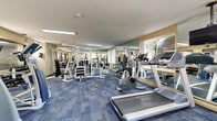 Located at 300 E Marshall St in West Chester, PA, this fitness center offers a well-equipped, single-room workout facility designed to support a full range of exercise routines. The space is finished with durable blue tile-pattern carpeting throughout and is illuminated by recessed fluorescent ceiling lights that keep the environment bright and functional. Floor-to-ceiling mirrors line the walls, visually expanding the room and allowing users to monitor their form during workouts. Natural light filters in through multiple windows, providing views of the surrounding greenery and adding to the open, energizing atmosphere. The cardio section is equipped with multiple treadmills and stationary bikes, as well as stair climber machines, catering to a variety of aerobic workout preferences. A ceiling-mounted television keeps users entertained during cardio sessions. The strength training area features a multi-station cable and weight machine system alongside a dedicated dumbbell rack stocked with a range of free weights and an adjustable bench. Glass-panel partitions and a glass entry door give the space a clean, modern aesthetic. The room's layout is thoughtfully organized to maximize usable floor space, ensuring smooth circulation between equipment stations while accommodating multiple users simultaneously.