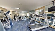 Located at 300 E Marshall St in West Chester, PA, this fitness center offers a well-equipped, single-room workout facility designed to support a full range of exercise routines. The space is finished with durable blue tile-pattern carpeting throughout and is illuminated by recessed fluorescent ceiling lights that keep the environment bright and functional. Floor-to-ceiling mirrors line the walls, visually expanding the room and allowing users to monitor their form during workouts. Natural light filters in through multiple windows, providing views of the surrounding greenery and adding to the open, energizing atmosphere. The cardio section is equipped with multiple treadmills and stationary bikes, as well as stair climber machines, catering to a variety of aerobic workout preferences. A ceiling-mounted television keeps users entertained during cardio sessions. The strength training area features a multi-station cable and weight machine system alongside a dedicated dumbbell rack stocked with a range of free weights and an adjustable bench. Glass-panel partitions and a glass entry door give the space a clean, modern aesthetic. The room's layout is thoughtfully organized to maximize usable floor space, ensuring smooth circulation between equipment stations while accommodating multiple users simultaneously.